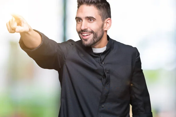 Young Christian priest over isolated background Pointing with finger surprised ahead, open mouth amazed expression, something in front