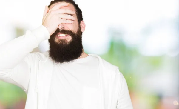 Young man with long hair and beard wearing sporty sweatshirt smiling and laughing with hand on face covering eyes for surprise. Blind concept.