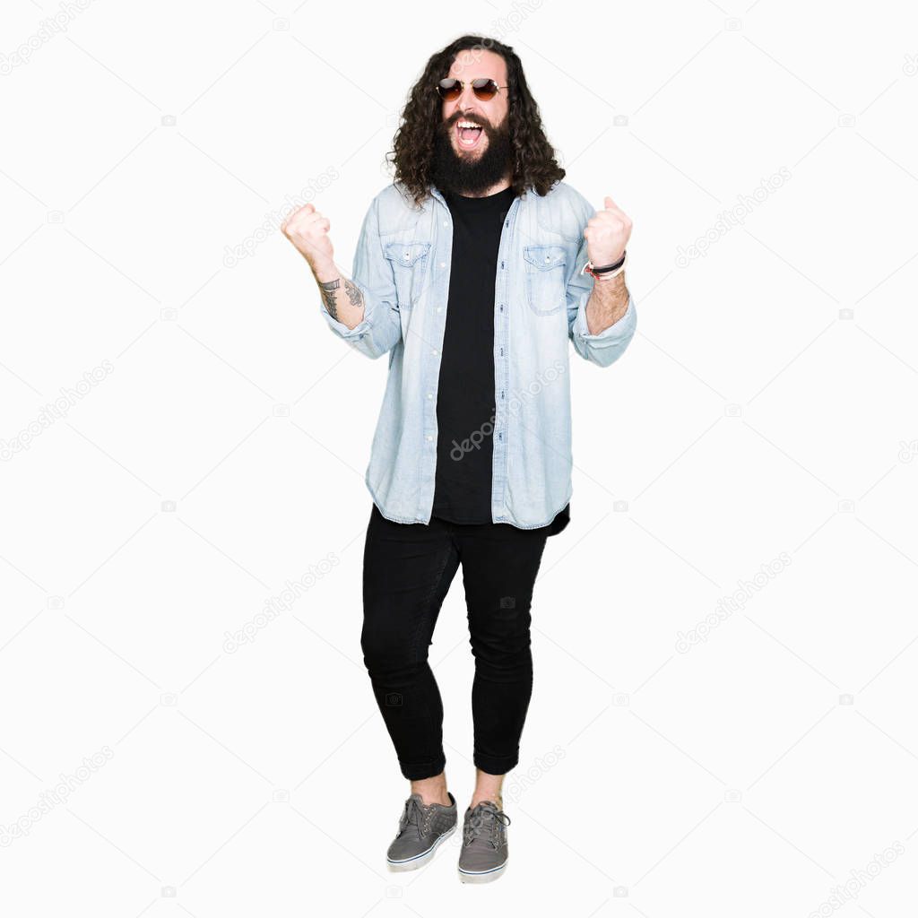 Young hipster man with long hair and beard wearing sunglasses very happy and excited doing winner gesture with arms raised, smiling and screaming for success. Celebration concept.