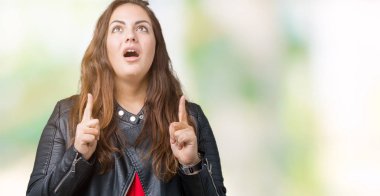 Artı boyutu genç kadın güzel izole arka plan üzerinde moda deri ceket hayret ve yukarıya bakıyor ve parmakları ile işaret ve silah kaldırdı şaşırttı.