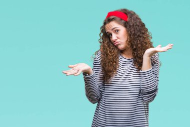 Güzel esmer kıvırcık saçlı Genç kız izole arka plan clueless ve şaşkın bir ifadeyle kollar ve eller kaldırdı üzerinde çizgili kazak giyiyor. Şüphe kavramı.