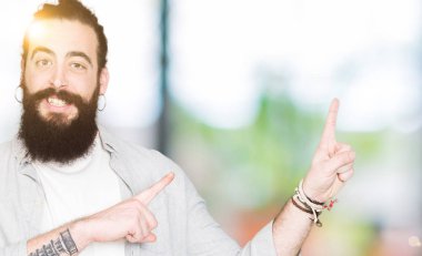 Uzun saç, sakal ve gülümseyen ve iki elleri ve parmakları ile tarafına işaret kamera bakarak küpe ile genç adam.