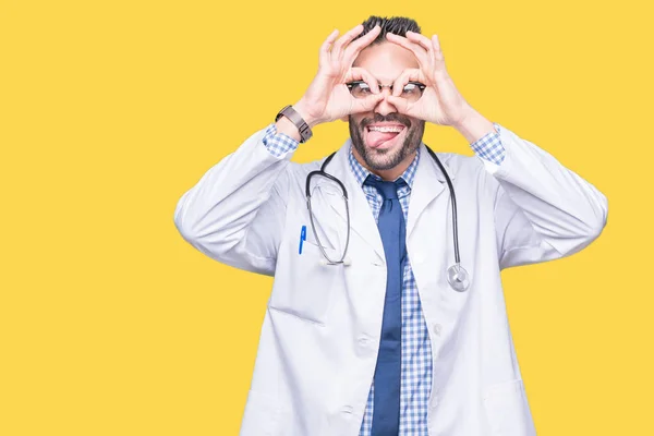 Handsome young doctor man over isolated background doing ok gesture like binoculars sticking tongue out, eyes looking through fingers. Crazy expression.