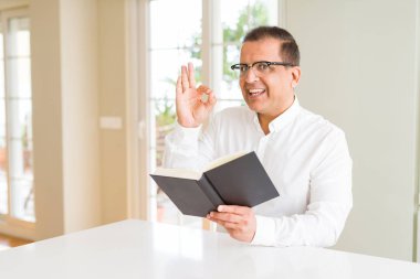 Evde kitap okurken orta yaş adam gözlüklü parmak, mükemmel sembolü ile ok işareti yapıyor