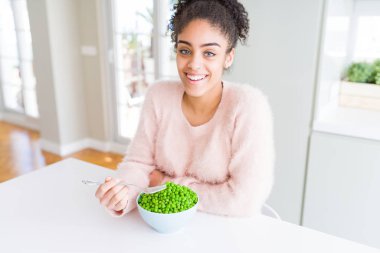 Genç Afrikalı Amerikalı kız mutlu bir yüz ayakta ve kendine güvenen bir gülümseme dişleri gösteren ile gülümseyen sağlıklı yeşil bezelye yeme