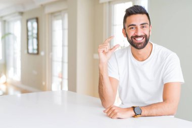 Yakışıklı İspanyol adam rahat beyaz t-shirt evde gülümseyen ve emin el ile boyutu işareti yaparken parmaklar ile görünümlü ve kamera bakıyor. Ölçü kavramı.
