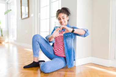 Afro saçlı güzel genç Afrikalı Amerikalı kadın yerde oturan aşk kalp sembolü ve elleri ile şekil gösteren gülümsüyor. Romantik konsept.