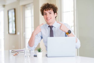 Genç iş adamı kendine güvenen gösterilen ve işaret parmakları diş ve ağız ile gülümseyen ofiste bilgisayar laptop ile çalışma. Sağlık kavramı.