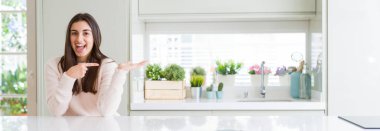 Geniş açı resim beyaz masaya oturan güzel genç kadının evde şaşırdım ve el ile sunulması ve parmak ile işaret ederken kameraya gülümseyen.