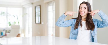 Evde beyaz masada oturan güzel genç kadının geniş açı resim, parmaklar ve ağız ile gösteren ve işaret emin gülümseyen. Sağlık konsepti.