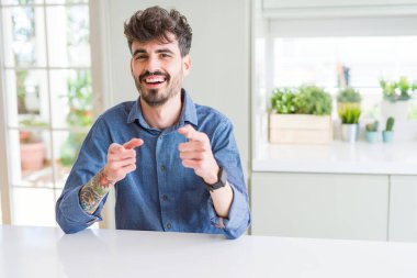 Beyaz masada oturan rahat gömlek giyen genç adam mutlu ve komik yüz ile kamera parmaklar işaret. İyi enerji ve Vibes.