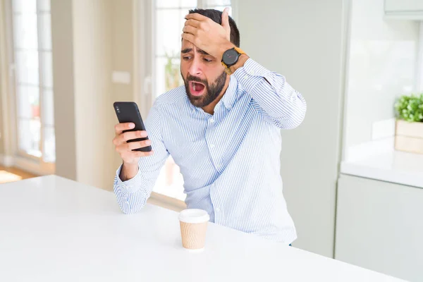 Handsome hispanic business man drinking coffee and using smartphone stressed with hand on head, shocked with shame and surprise face, angry and frustrated. Fear and upset for mistake.