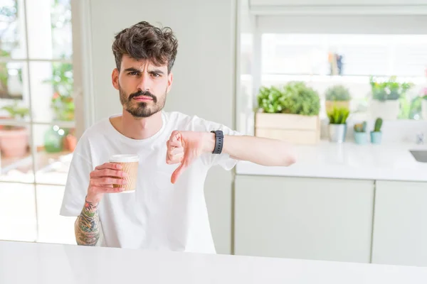 Young man drinking a coffee in a paper cup in the morning with angry face, negative sign showing dislike with thumbs down, rejection concept