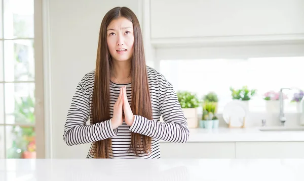 Beautiful Asian woman wearing stripes sweater begging and praying with hands together with hope expression on face very emotional and worried. Asking for forgiveness. Religion concept.