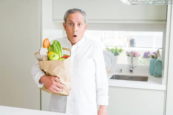 Handsome senior man holding a paper bag of fresh groceries at the kitchen scared in shock with a surprise face, afraid and excited with fear expression