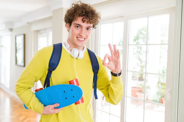 ヘッドフォンと優秀なシンボルの指で サインをしているスケート ボードを持ってのバックパックを身に着けている若い学生男 — ストック写真