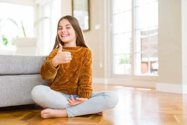 Evde yerde oturan güzel genç kız çocuk el ile jest kadar mutlu başparmak yapıyor. Başarı göstererek kameraya bakarak ifadeyi onaylama.