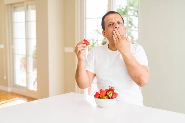 Middle age man eating strawberries at home cover mouth with hand shocked with shame for mistake, expression of fear, scared in silence, secret concept