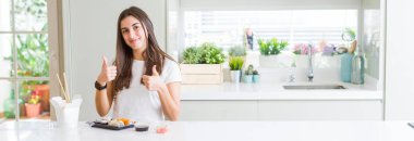 Güzel genç kadının geniş açı resmi teslim başarı işareti ile olumlu jest yaparak Asya Suşi yeme el, gülümseyen ve mutlu kadar başparmak. Neşeli ifade ile kamera bakarak, kazanan jest.