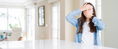 Evde beyaz masada oturan güzel genç kadının geniş açı resmi eller ile gözler ve ağız kapsayan, sürpriz ve şok. Duyguyu gizleme