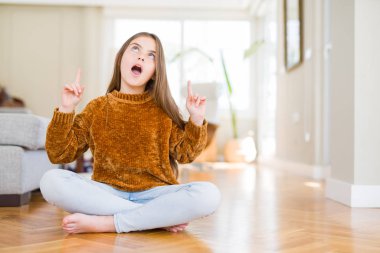 Evde yerde oturan güzel genç kız çocuk şaşırır ve görünümlü ve parmaklar ve yükseltilmiş kollar ile işaret şaşırttı.