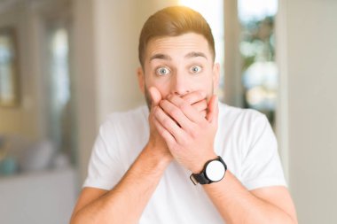 Evde rahat beyaz t-shirt giyen genç yakışıklı adam yanlışlık için eller ile ağız kapsayan şok. Gizli konsept.