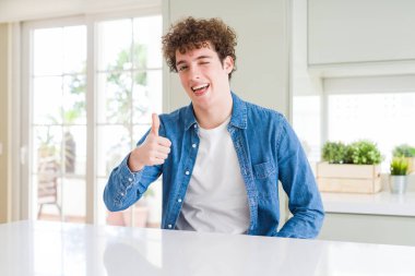 Evde rahat denim ceket giyen genç yakışıklı adam hareketi el ile mutlu Yaşasın yapıyor. Başarı gösteren kameraya bakarak ifade onaylama.