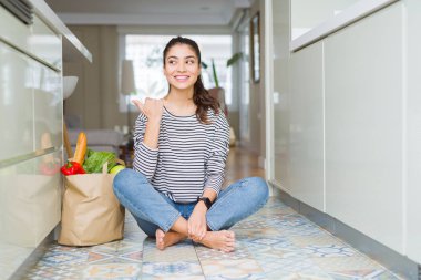 Genç kadın, mutlu yüz görünümlü ve başparmak yukarı ile yan işaret ile gülümseyen taze bakkallarla dolu bir kağıt çanta ile mutfak katında oturan.