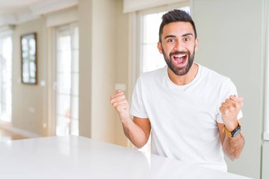 Evde yakışıklı İspanyol adam Casual beyaz t-shirt sürpriz kutluyor ve silah yükseltilmiş ve açık gözleri ile başarı için hayran. Kazanan kavramı.