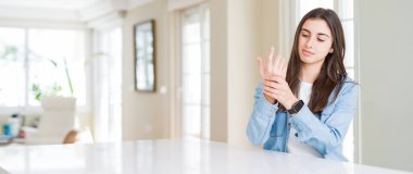 Evde beyaz masada oturan güzel genç kadının geniş açı resmi ellerde ve parmaklarda ağrı acı, artrit iltihabı