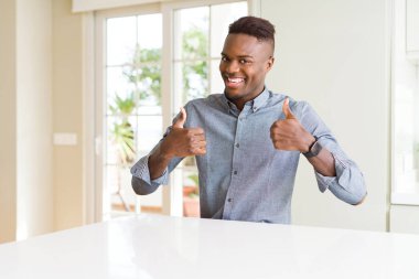 Afro-Amerikan yakışıklı beyaz masa başarı işareti el ile pozitif jest yapıyor başparmak gülümseyen ve mutlu. Neşeli bir ifadeyle, kazanan jest kameraya bakıyor.