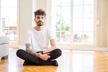 Genç adam yüzünde ciddi bir ifade ile evde rahat oturma çalışıyor. Basit ve doğal kamera bakıyor.