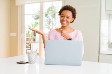 Hayret ve el ile sunulması ve parmak ile işaret ederken kameraya gülümseyen bilgisayar dizüstü kullanarak çalışan genç Afro-Amerikan kadın.