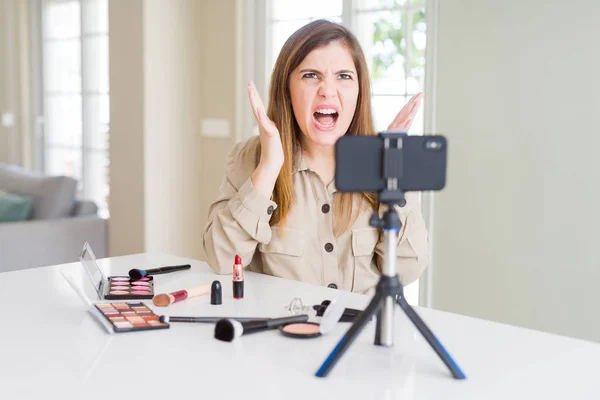 Beautiful young influencer woman recording make up video tutorial crazy and mad shouting and yelling with aggressive expression and arms raised. Frustration concept.