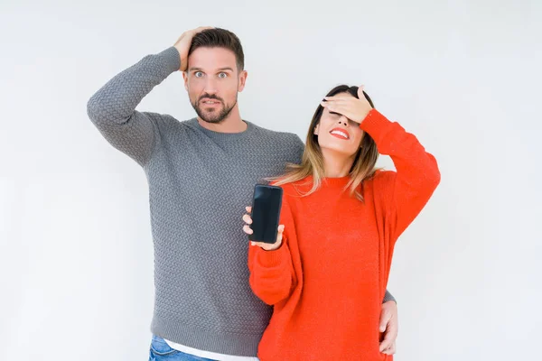 Young couple showing smartphone screen over isolated background stressed with hand on head, shocked with shame and surprise face, angry and frustrated. Fear and upset for mistake.