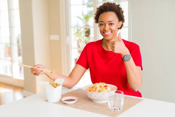 Young african american woman with afro hair eating asian food at home happy with big smile doing ok sign, thumb up with fingers, excellent sign