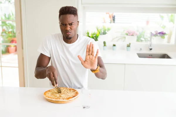 African american man eating cheese pizza at home with open hand doing stop sign with serious and confident expression, defense gesture