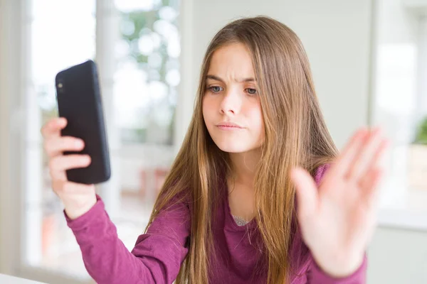 美丽的年轻女孩孩子发送的消息使用智能手机与开放的手做停止标志与严肃和自信的表达 防御手势 — 图库照片