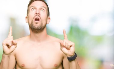 Çıplak göğüs gösteren yakışıklı gömleksiz adam hayret ve bakmak ve parmaklar ve yükseltilmiş silah ile işaret şaşırttı.