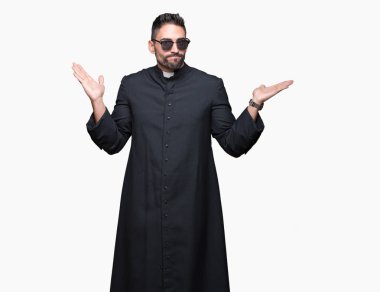 Genç Hıristiyan rahip giyen güneş gözlüğü izole arka plan clueless ve şaşkın bir ifadeyle kollar ve eller ortaya çıkar üzerinden. Şüphe kavramı.