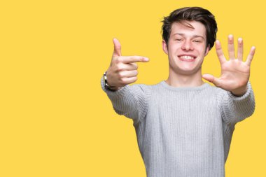 Gösterilen arka plan üzerinde kış kazak giyen genç yakışıklı adam izole ve parmakları ile işaret kendinden emin ve mutlu gülümseyerek süre yedi numara.