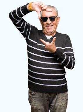 Mutlu yüz ile eller ve parmaklar ile çerçeve yapma gülümseyen kış kazak ve güneş gözlüğü giyen yakışıklı kıdemli adam. Yaratıcılık ve Fotoğrafçılık konsepti.