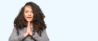 Kıvırcık saçlı Genç güzel kadın, el ile birlikte AF gülümseyen emin gülümsüyor isteyen dua gri kazak giyiyor.