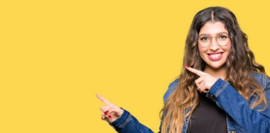 Gözlük ve gülümseyen ve iki elleri ve parmakları ile tarafına işaret kamera bakarak denim ceket giyen genç güzel kadın.