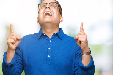 Orta yaş Arap adam izole arka plan üzerinde gözlük hayrete ve yukarıya bakıyor ve parmakları ile işaret ve silah kaldırdı şaşırttı.