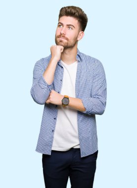 Genç yakışıklı sarışın adam el çenesi ile rahat gömlek giyiyor soru hakkında düşünme, dalgın ifade. Düşünceli bir yüz ile gülümsüyor. Şüphe kavramı.