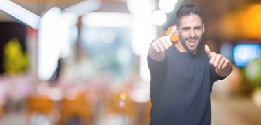 El ile pozitif jest yapıyor onaylama izole arka plan üzerinde kazak giyen genç yakışıklı adam başparmak gülümseyen ve başarı için mutlu. Kamera, kazanan jest arıyorsunuz.