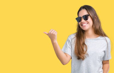 Genç güzel esmer kadın arıyorum ve başparmak yanında yukarıyı mutlu yüz ile gülümseyen izole arka plan üzerinde güneş gözlüğü takıyor.