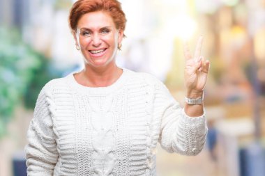 Mutlu yüz zafer işareti yaparken kameraya göz kırpıyor ile gülümseyen izole arka plan üzerinde kış kazak giyen Atrractive üst düzey beyaz Kızıl saçlı kadın. İki numaralı.