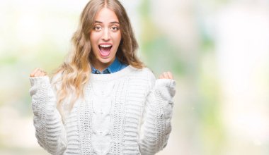 İzole arka plan üzerinde kutluyor kış kazak giyen güzel genç sarışın kadın şaşırttı ve başarı için kaldırdı kollarını açarak hayret ve gözleri açık. Kazanan kavramı.